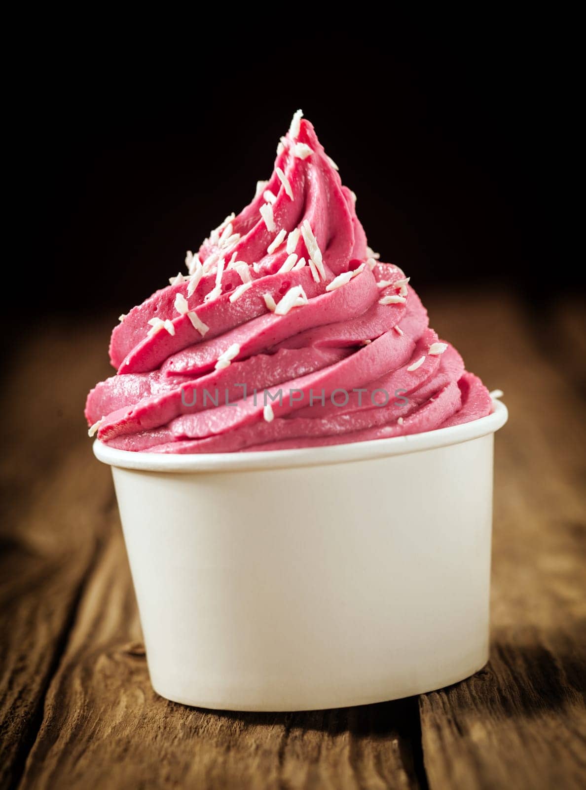 Twirl of delicious berry ice cream or frozen yoghurt in a plastic takeaway tub sprinkled with desiccated coconut against a dark background , close up side view
