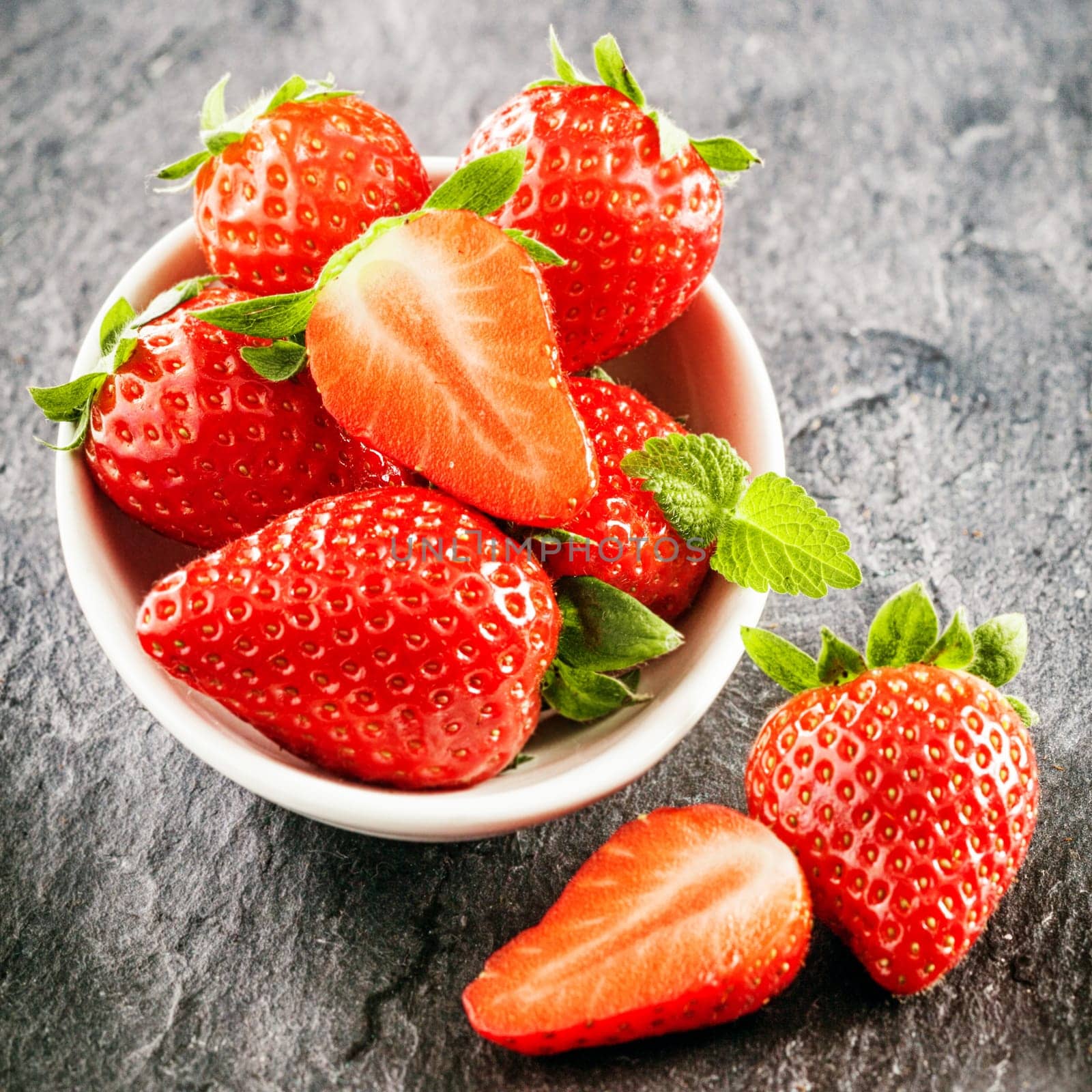 Fresh juicy farm fresh strawberries with attached green stalks served in a bowl on grey slate in square format