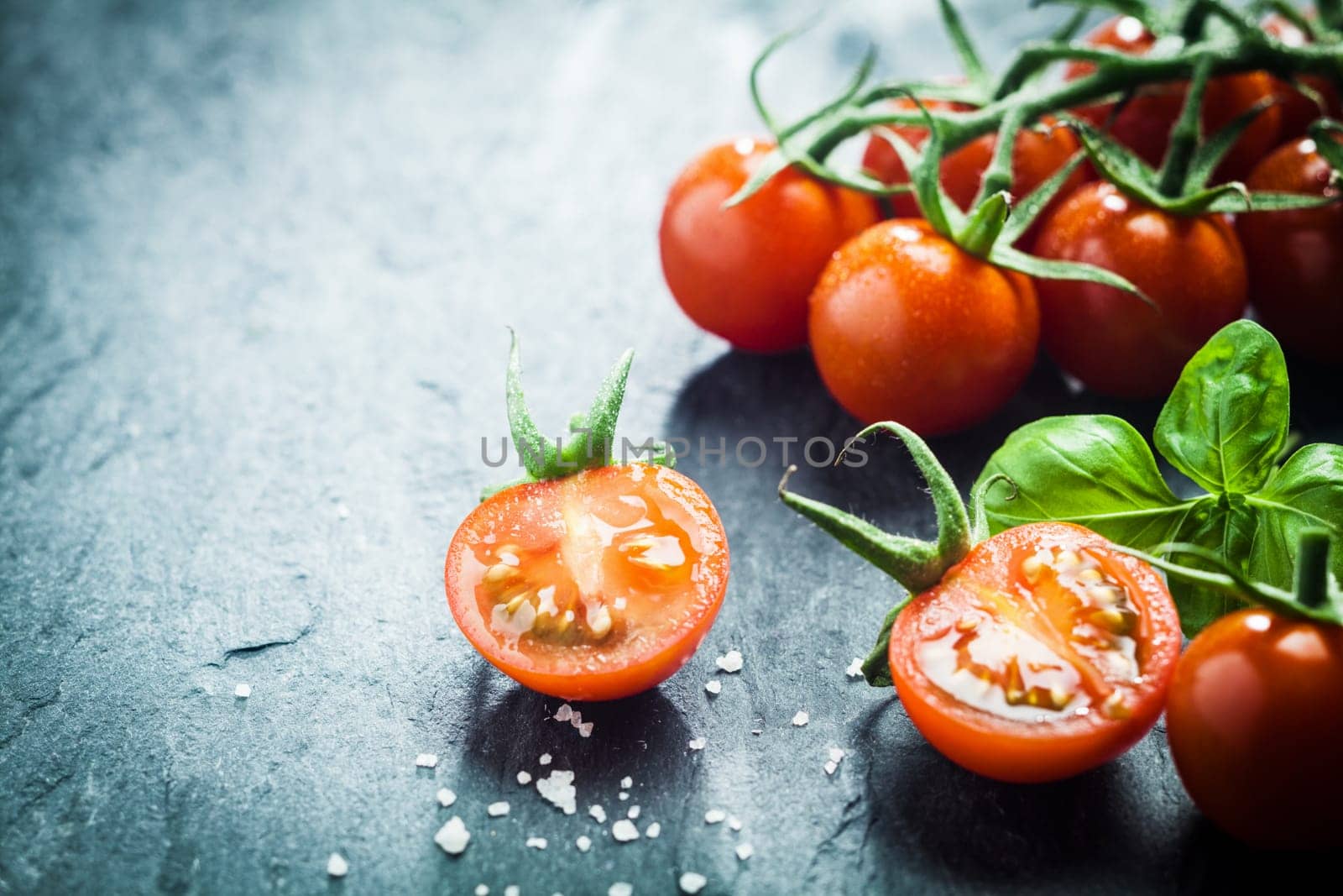 Fresh grape tomatoes with basil and coarse salt for use as cooking ingredients with a halved tomato in the foreground with copyspace