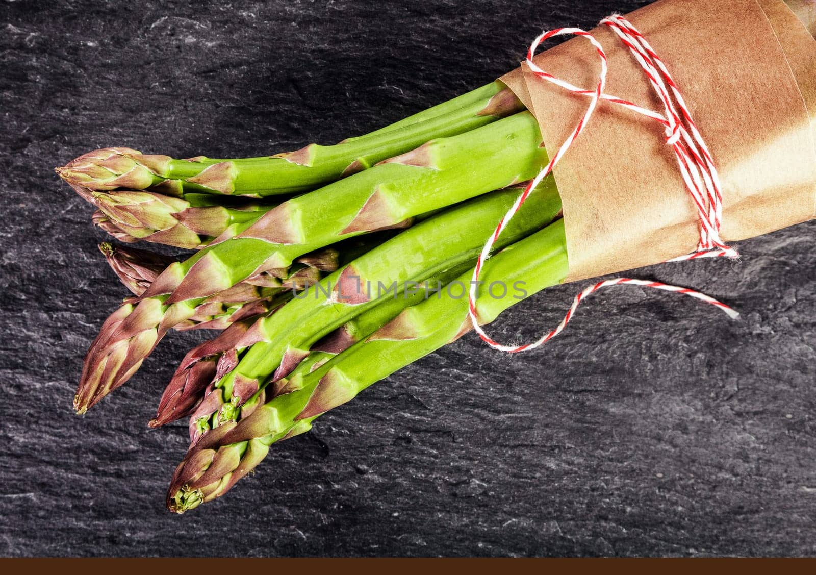 Fresh green asparagus shoots tied in a bundle with brown paper and string, close up overhead view on dark slate with copyspace