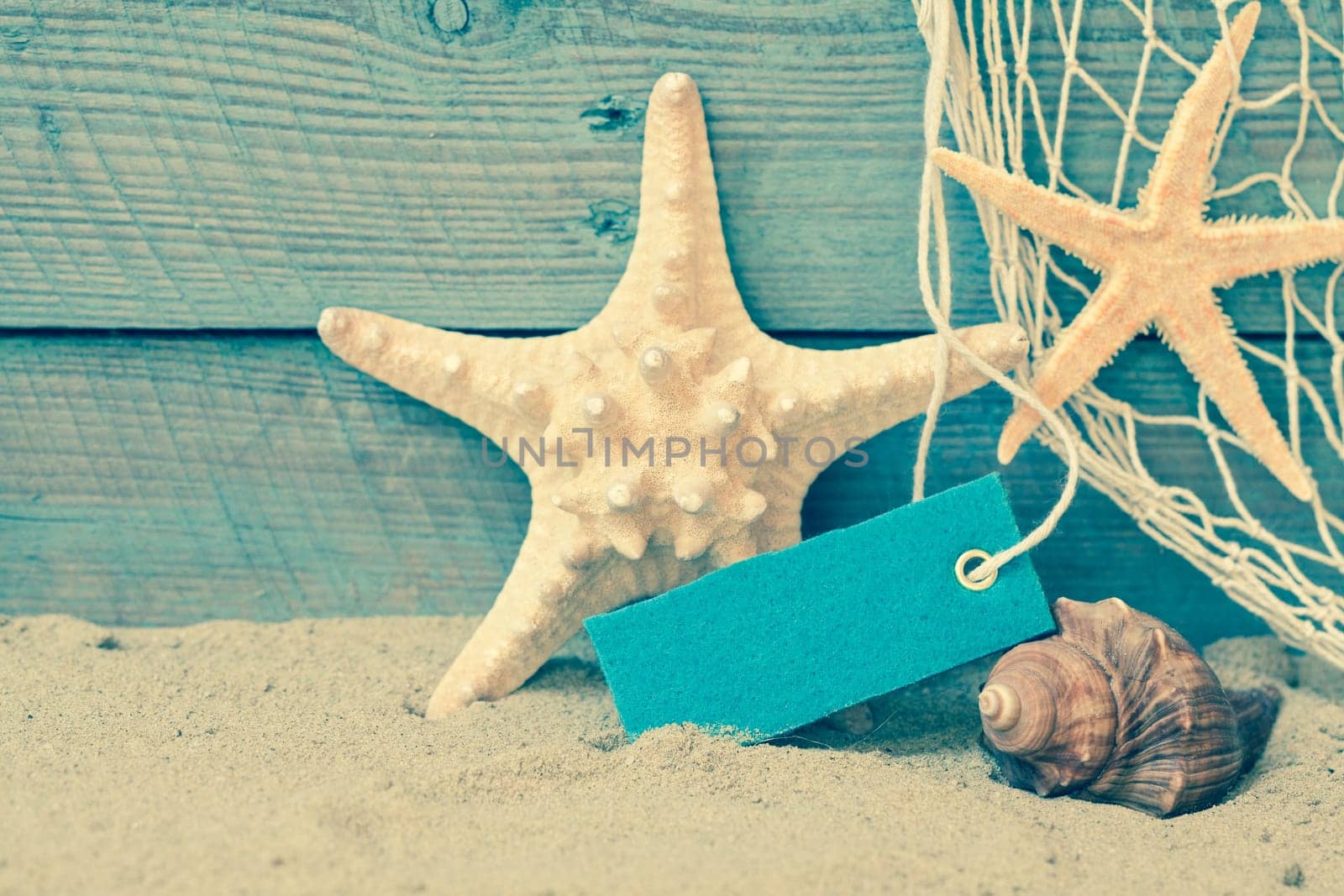 Toned retro nautical background with starfish and a seashell on beach sand against rustic blue wooden boards with a blank label or tag with copyspace and a decorative fishing net