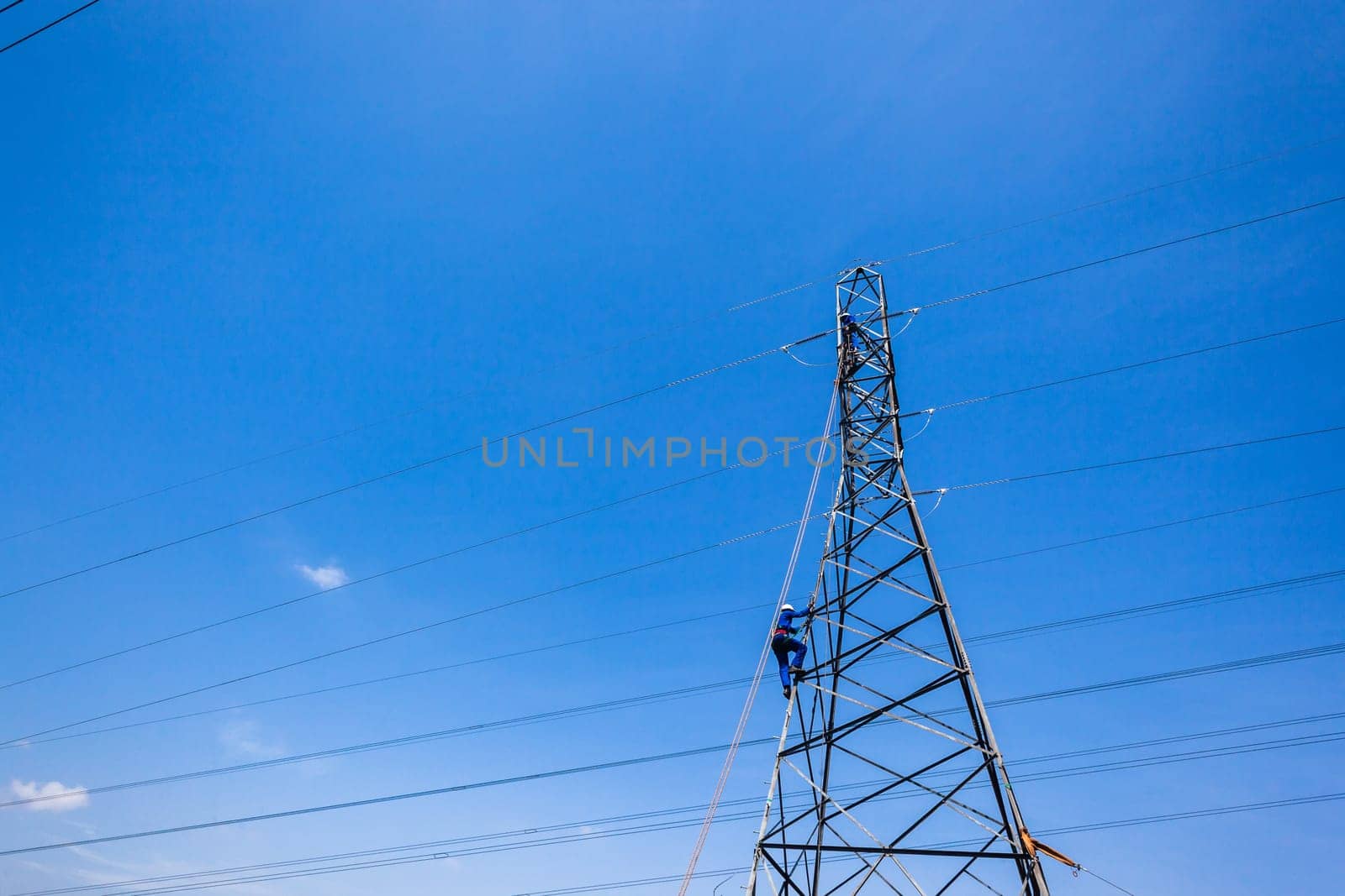 Electricians climb steel electrical tower for maintenance repairs on high powered electrical cables in blue sky.