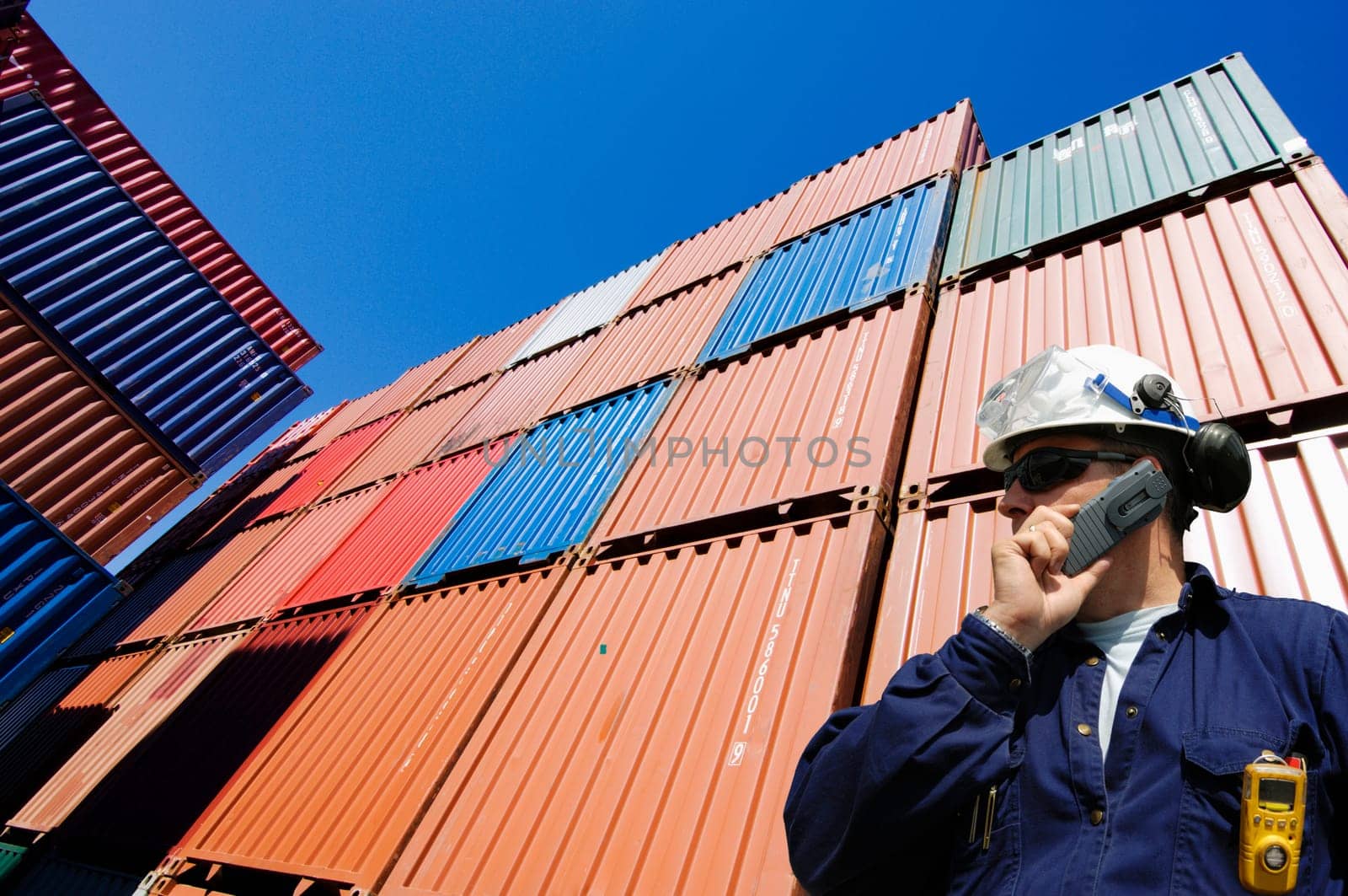 port and dock worker talking in phone with stacks of cargo containers in the background