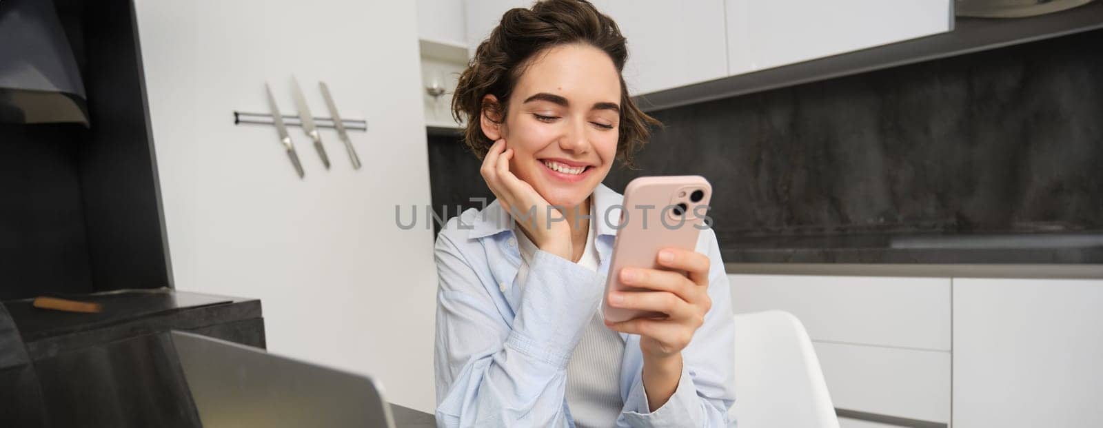 Image of young woman using her smartphone at home. Girl sits with mobile phone in kitchen and smiles by Benzoix