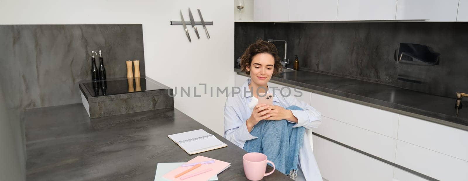 Young woman having break from work, sitting in kitchen and using smartphone, girl studies at home, sends a message on mobile phone.