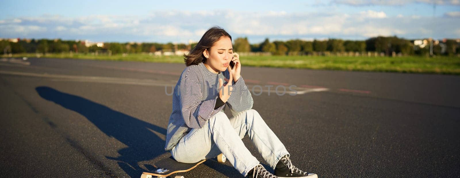 Portrait of asian girl sits on her longboard, skating on skateboard and talking on mobile phone, having conflict during telephone conversation, arguing with concerned face.