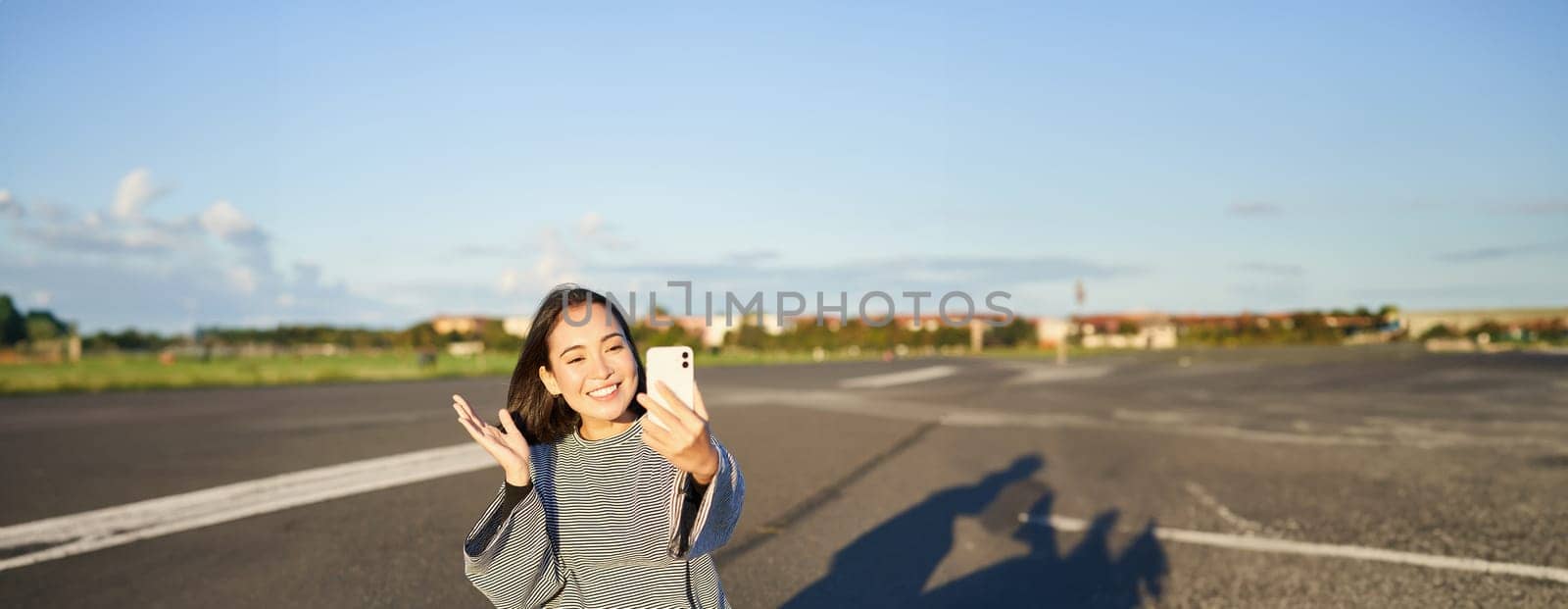 Happy teen asian girl takes selfie, video chats on smartphone app while sits on skateboard, skater records vlog while sits on longboard.