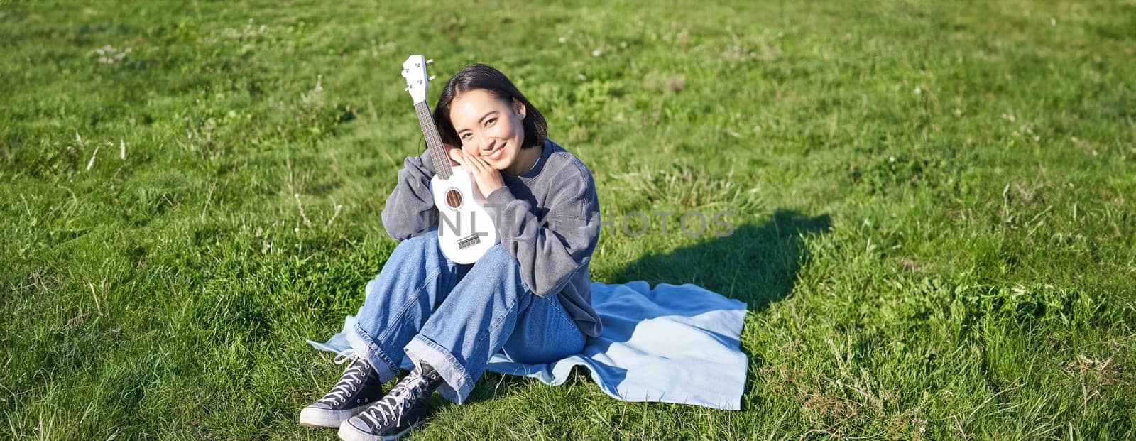 Cute romantic asian girl sitting with her instrument in park, hugging ukulele and smiling, resting outdoors by Benzoix