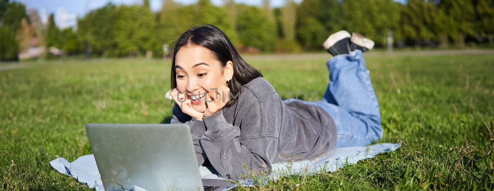 Portrait of beautiful girl lying in park and looking with surprised face at her laptop, watching videos, chatting with friends while relaxing outdoors.