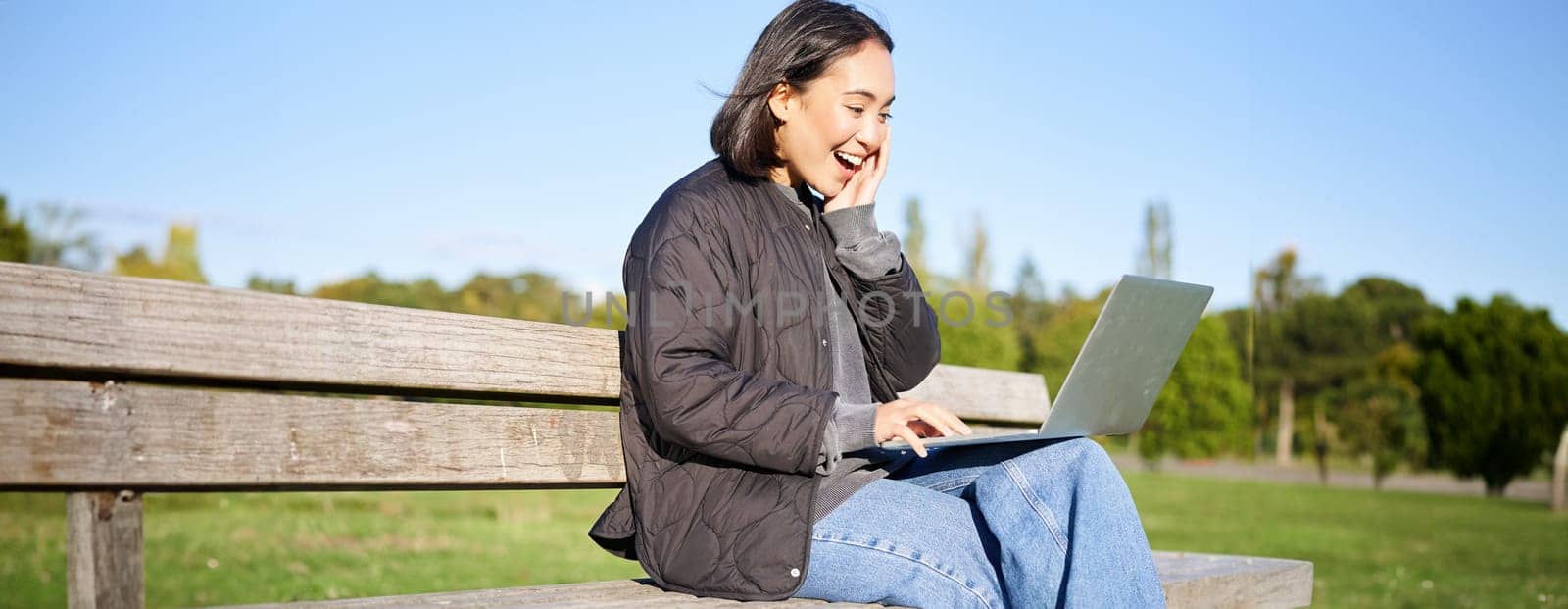 Portrait of smiling asian girl sits on bench in park, talks to friend online via laptop, video chats, using computer to study on remote.