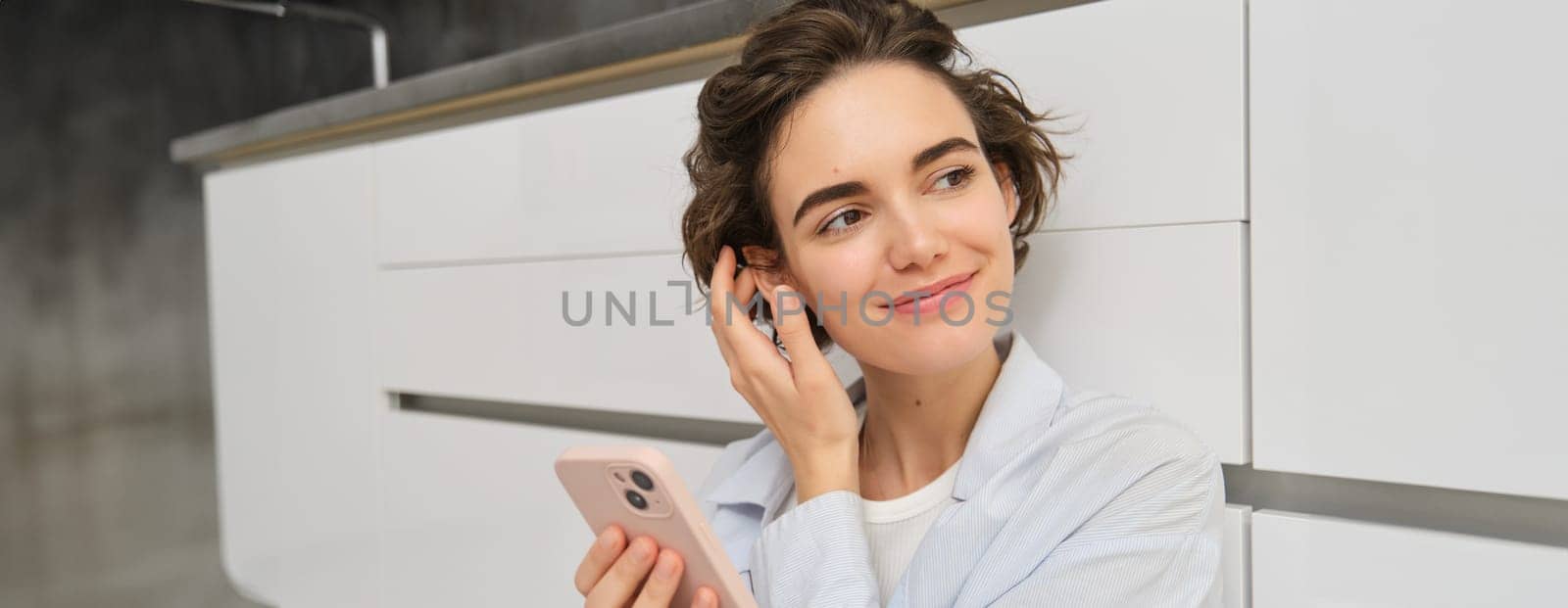 Indoor shot of happy girl with smartphone, sits on floor at home and uses mobile phone, watches video in app, chats with friends in applicatiion by Benzoix