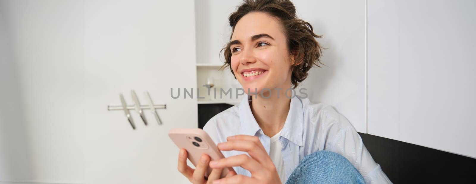 Portrait of smiling beautiful woman, sitting in kitchen with smartphone, looking happy at camera, spending time at home, using mobile phone app by Benzoix