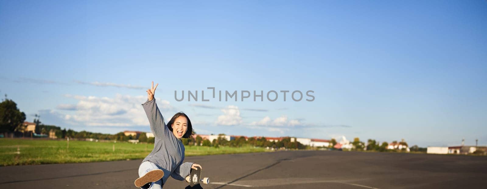 Vertical shot of happy asian skater girl, jumping, standing with skateboard and smiling. Woman skating on longboard and having fun.