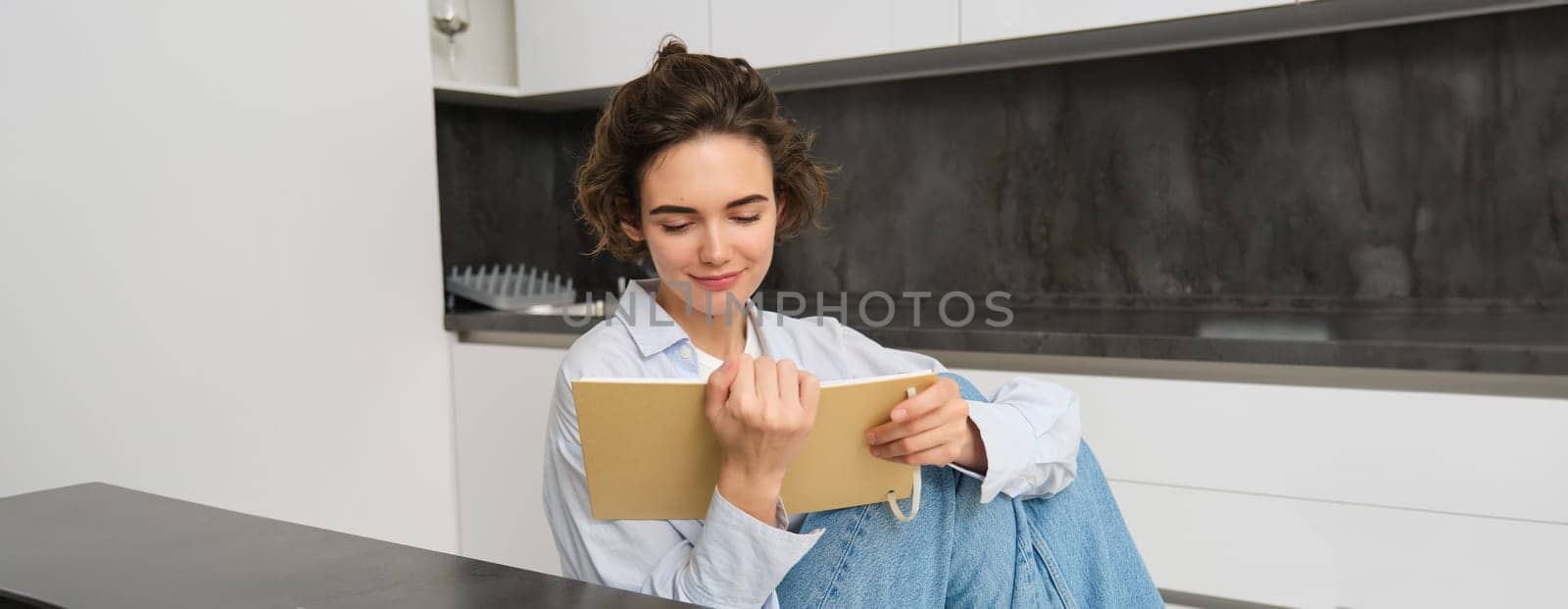 Young woman sitting in kitchen, reading her notes in notebook. Smiling girl at home, doing homework, studying.