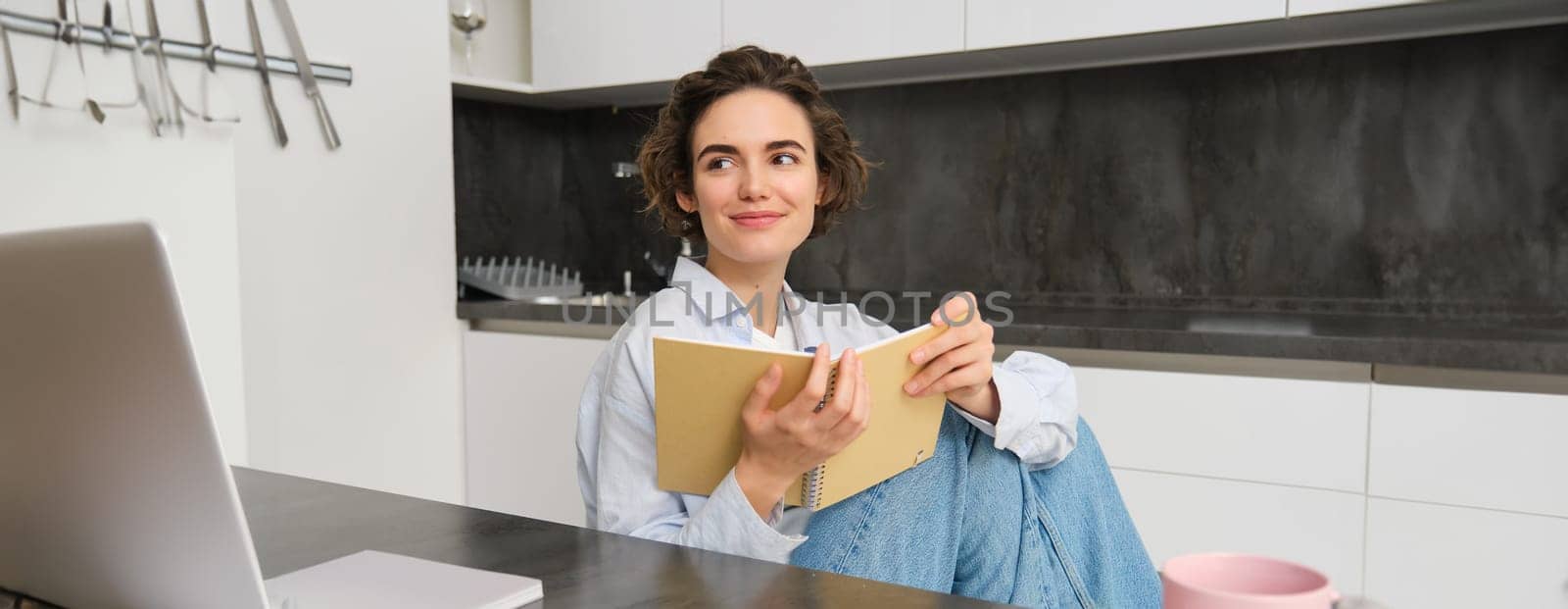 Young woman sitting in kitchen, reading her notes in notebook. Smiling girl at home, doing homework, studying.