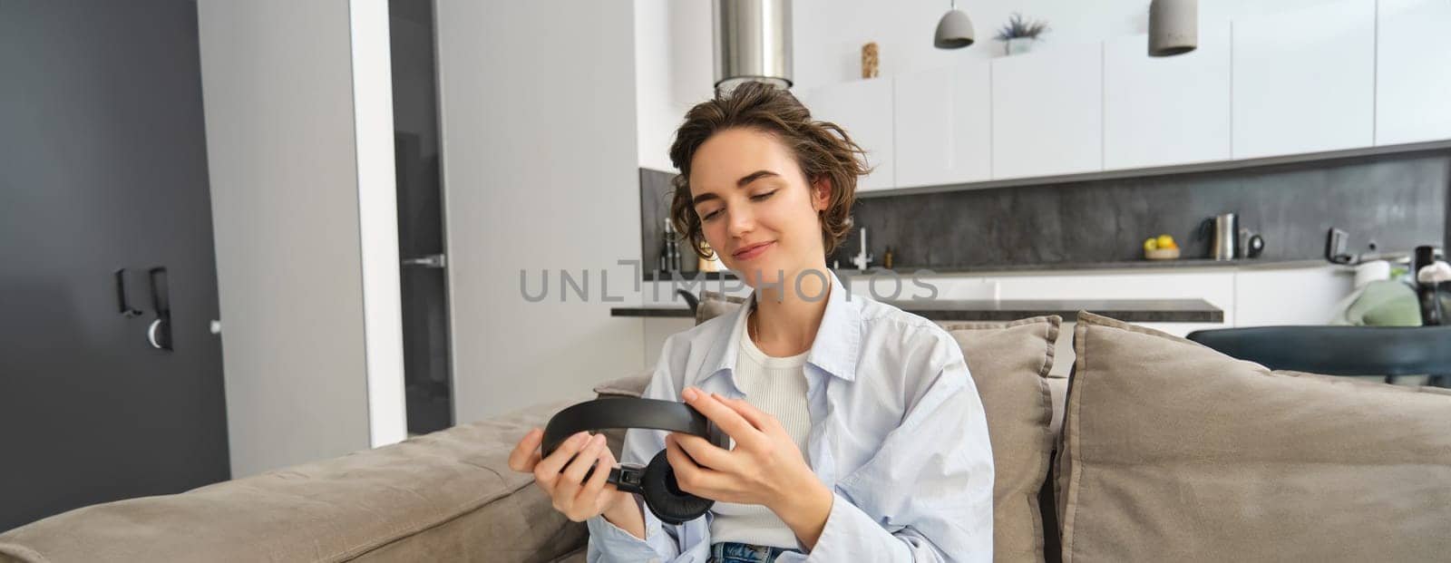 Portrait of beautiful, smiling woman looking at her headphones, listens to podcast or music while sits at home on sofa and relaxed on weekend.