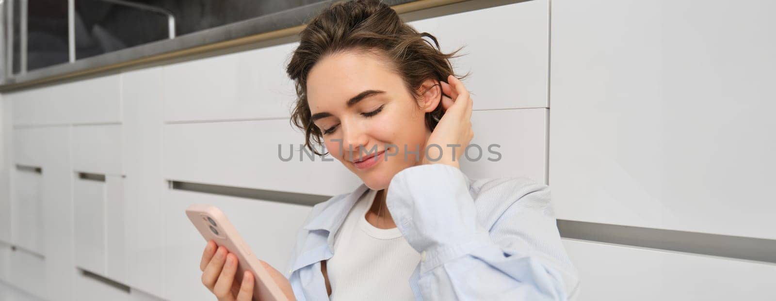 Portrait of cute smiling woman, sitting on floor at home with crossed legs, looks at mobile phone, chats on smartphone app, watches videos in application.