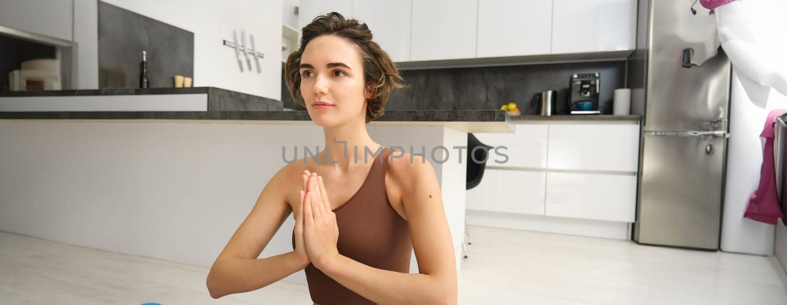 Smiling girl athlete, sportswoman doing yoga at home in activewear, sitting on yoga rubber mat in lotus pose, meditating, practice mindfulness exercises.