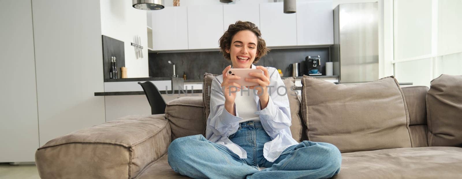 Portrait of young girl gamer, plays mobile video game on smartphone, sits on sofa with excited face expression by Benzoix