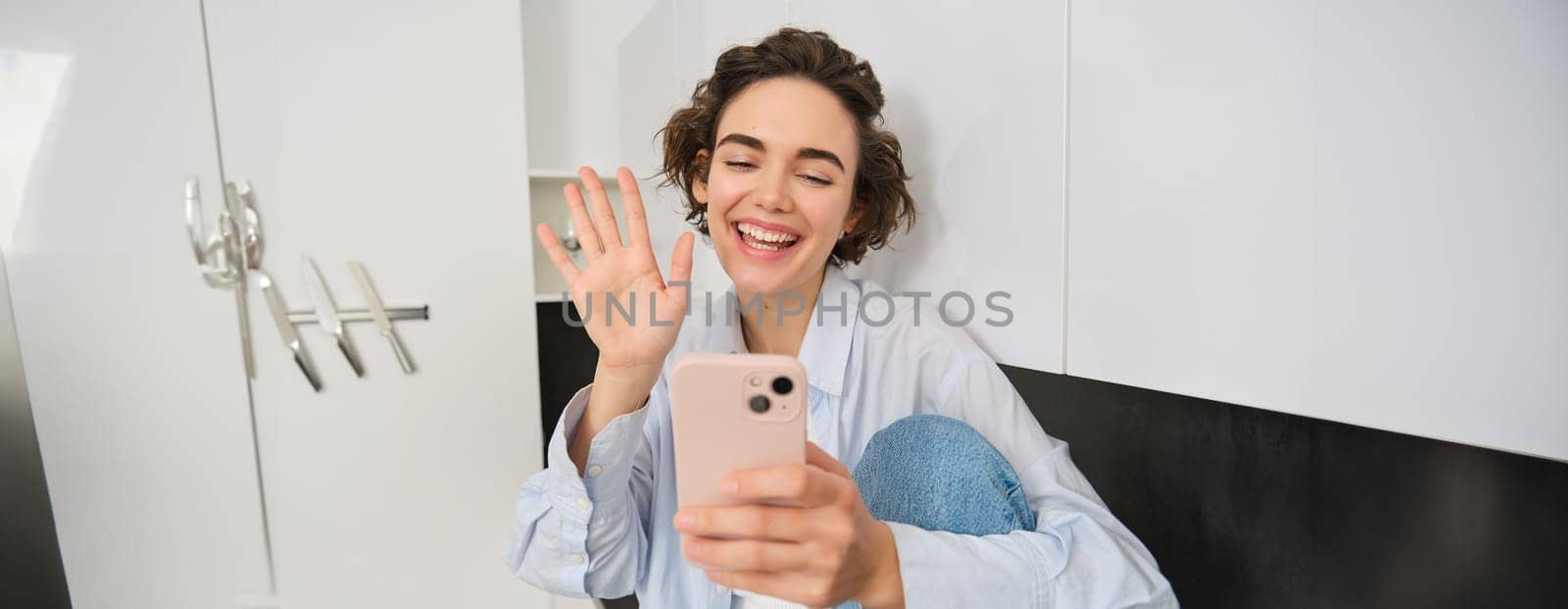 Portrait of smiling friendly woman, looking at mobile phone, waves hand at smartphone camera, video chats, connects to online conversation, sits at home and talks with someone.