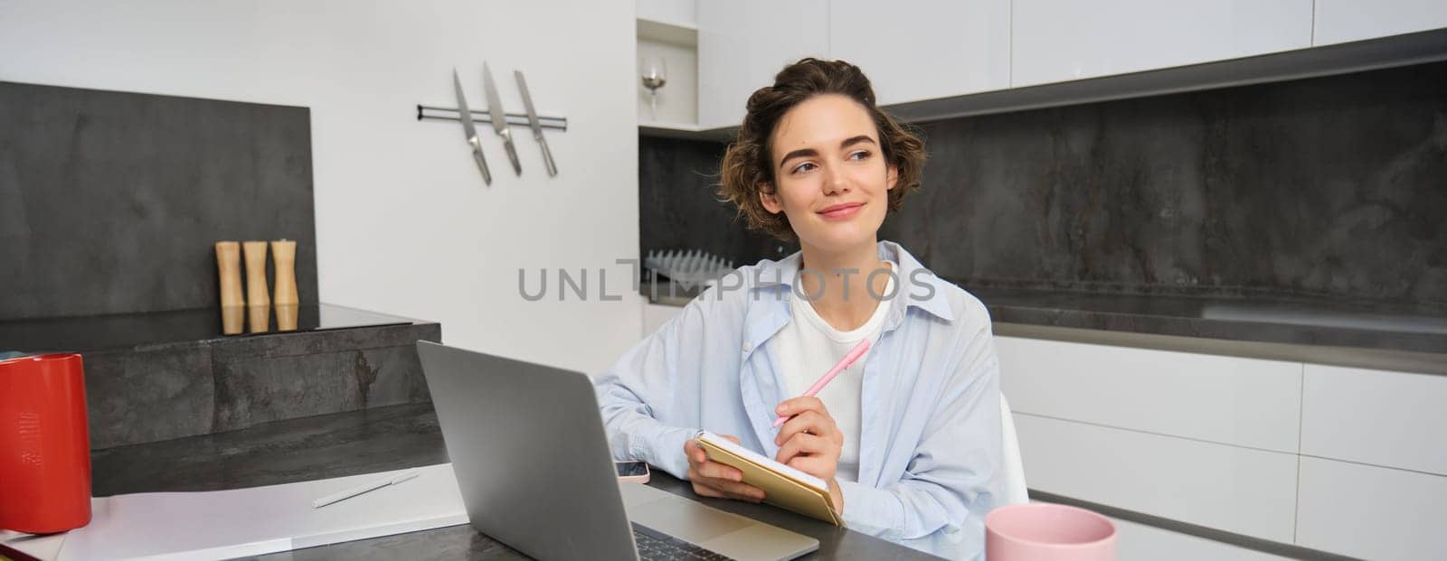 Smiling creative woman, holds pen and notebook, looks thoughtful, works from home with laptop in kitchen, thinking.