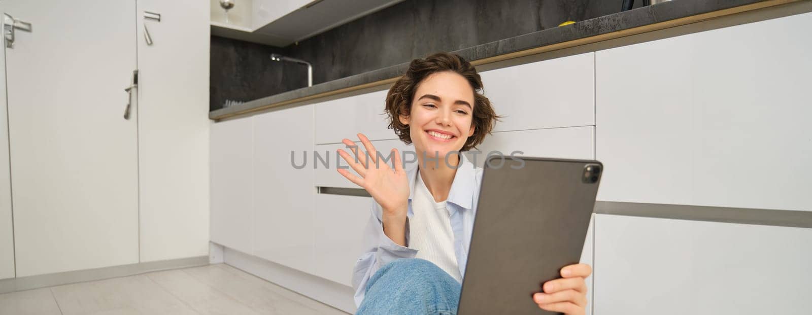 Portrait of happy young woman connects to video chat, waves at digital tablet and smiling, sits at home by Benzoix