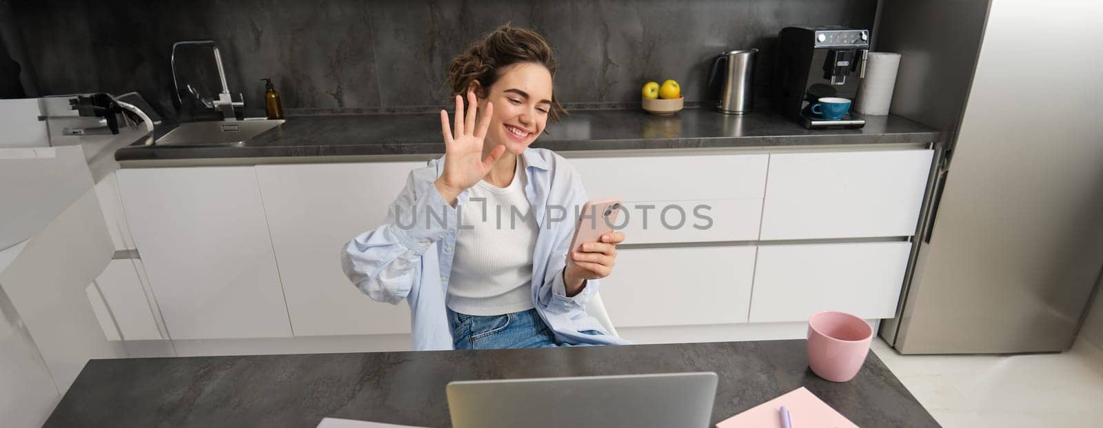 Young friendly woman, waves hand at laptop camera, video chats, holds smartphone, has conversation online.