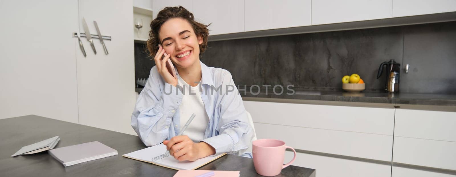 Portrait of candid, smiling young woman, talking on mobile phone, laughing during conversation on smartphone, writing down, making notes in notebook, sitting in kitchen at home.