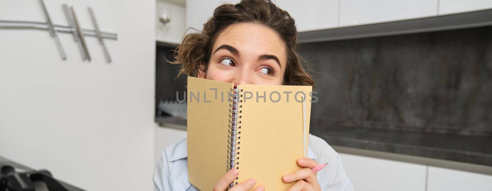 Portrait of cute brunette girl, looks away, hides her face behind planner, notebook, gets creative and writes in her journal.
