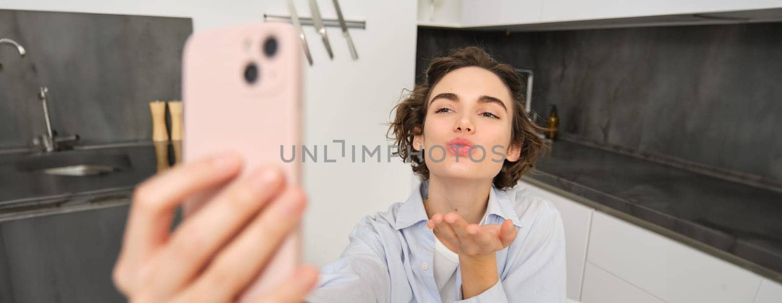 Portrait of beautiful young woman, taking selfie on mobile phone at home, poses for photo with smartphone by Benzoix