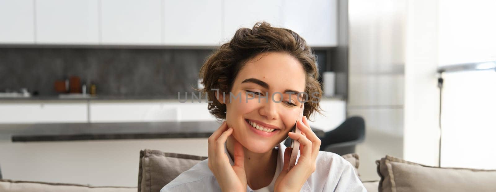 Close up portrait of happy brunette girl, talks on mobile phone, chats on smartphone, calls someone while spending time at home on sofa.