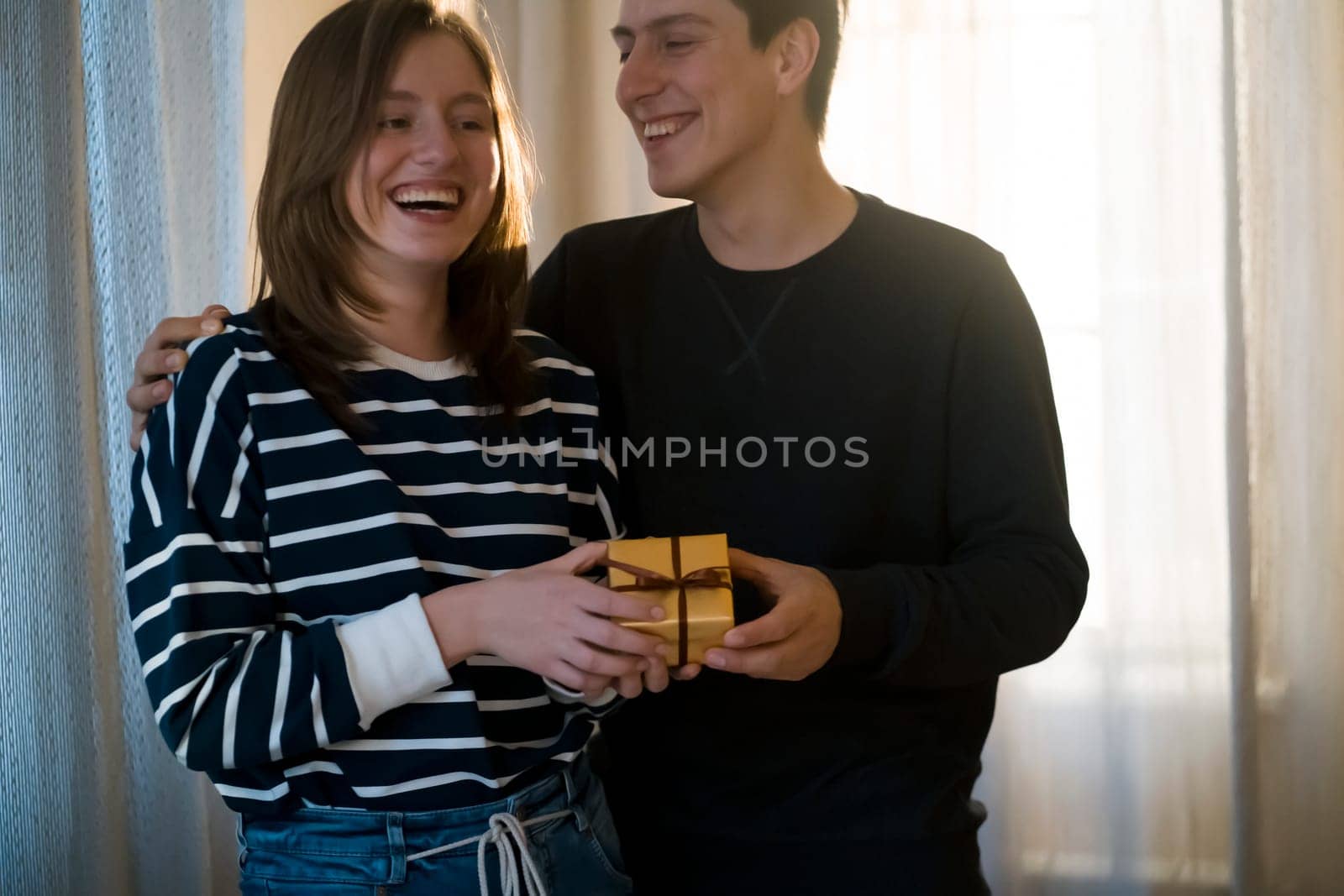 Young man and woman laugh and have a good time together in their cozy home, feel unity. Couple celebrating holidays, valentine's day, anniversary, new year or birthday and give each other cute gifts.