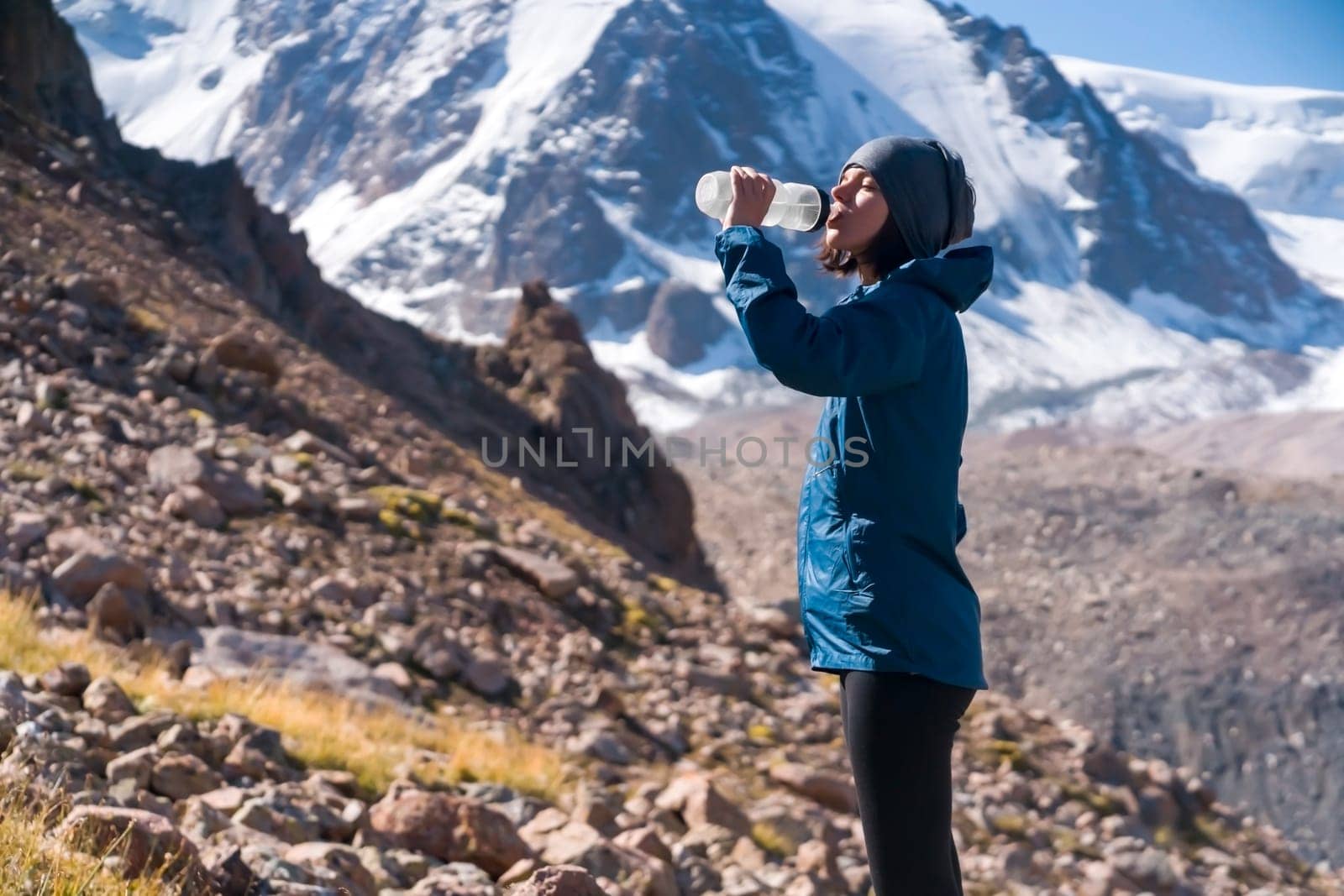 Young sports girl stands on the backdrop of a stunning view with snow-capped mountains and drinks clear water from a bottle. Woman is trail running, training, climbing and drinking isotonic.