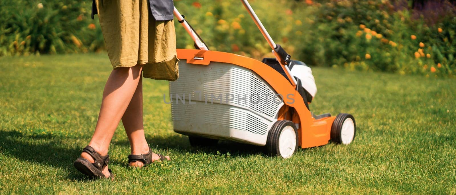 A young girl is mowing a lawn in the backyard with an orange lawn mower. A woman gardener is trimming grass with the grass cutter. A lawnmower is cutting a lawn on a summer sunny day.