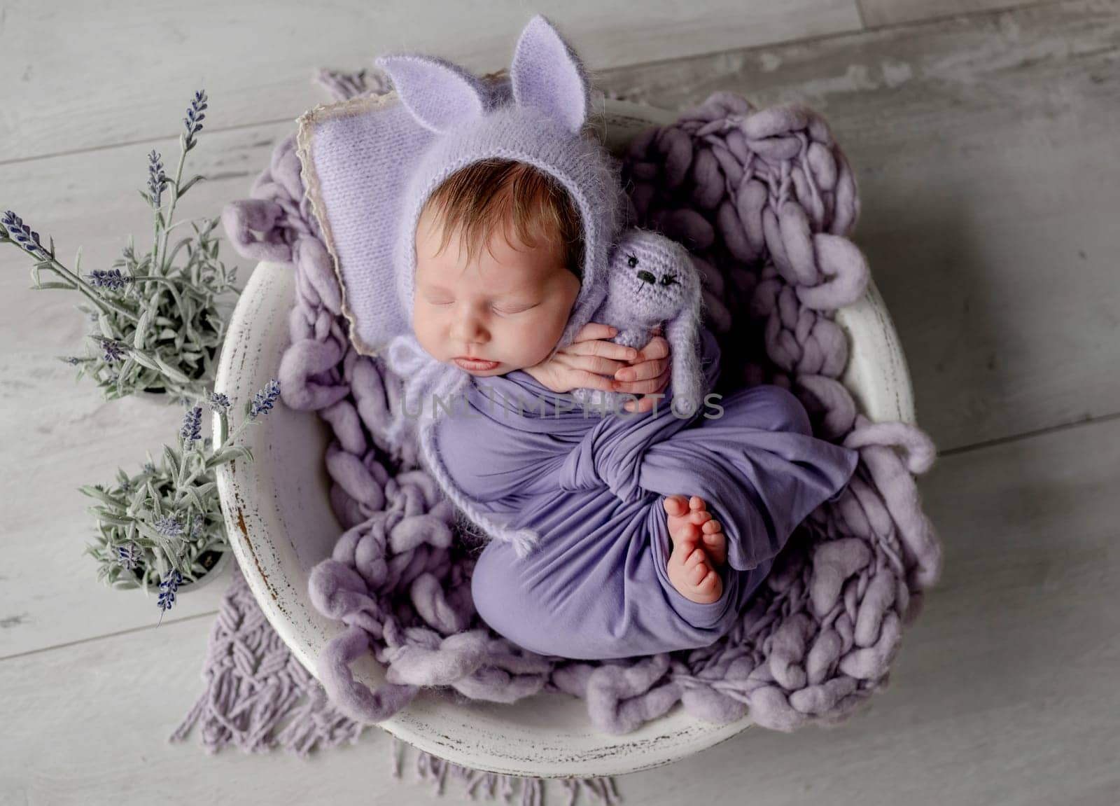 Newborn baby girl wearing knitted hat with rabbit ears holding bunny toy and looking at camera. Infant child kid lying swaddled in purple fabric studio portrait