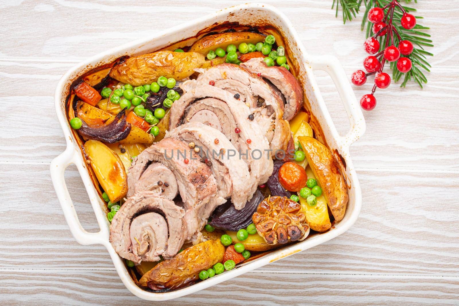 Festive Christmas rolled sliced pork roasted in white casserole dish with potatoes, vegetables and herbs on rustic white wooden background top view. Baked pork roll with vegetables for Xmas dinner by its_al_dente