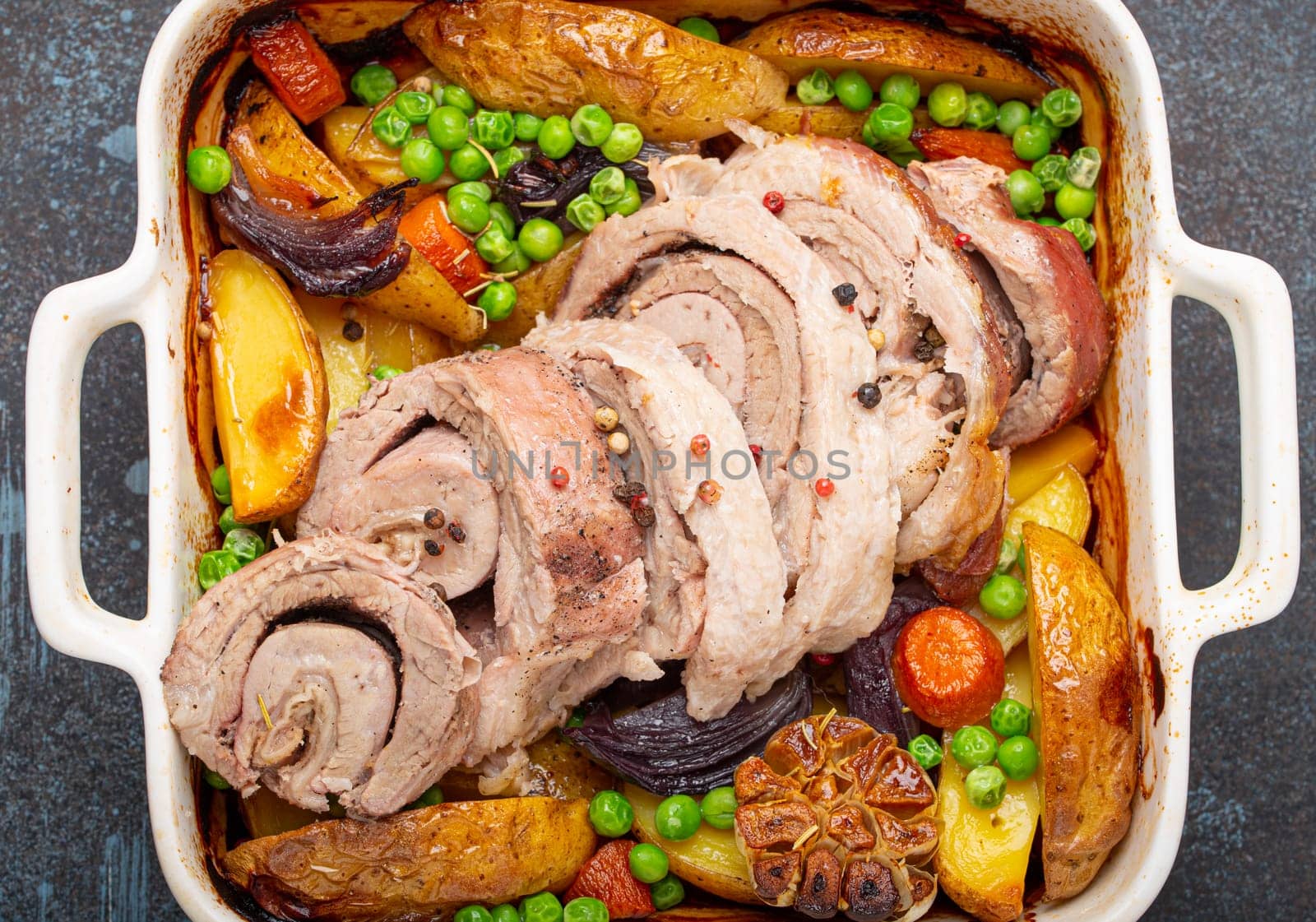 Rolled sliced pork roasted in white casserole dish with potatoes, vegetables and herbs on blue dark concrete rustic background top view. Baked pork roll with vegetables for dinner.