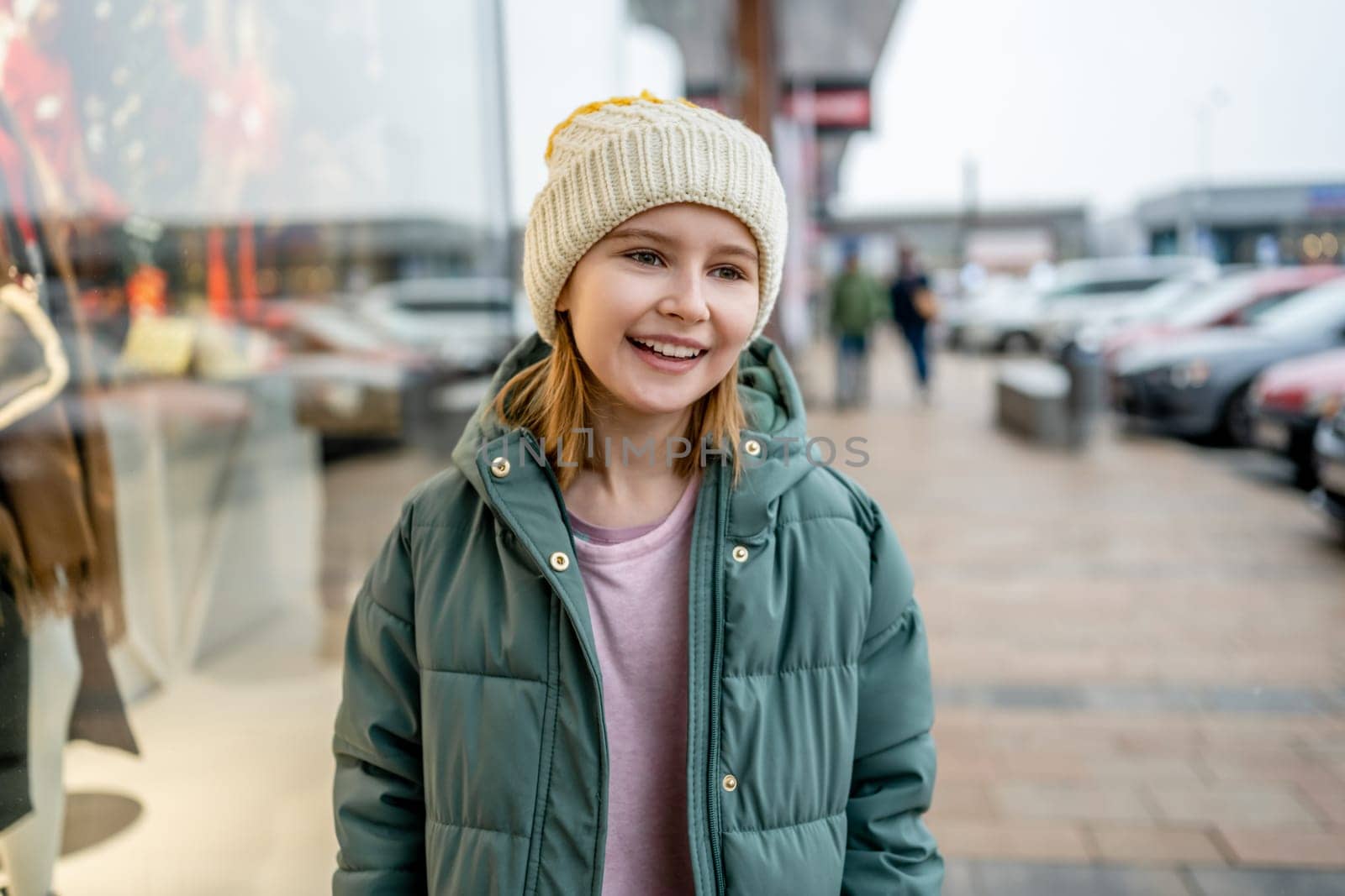 Preteen girl smiling at street portrait in city with blurred background. Cute female child kid wearing hat outdoors at autumn time