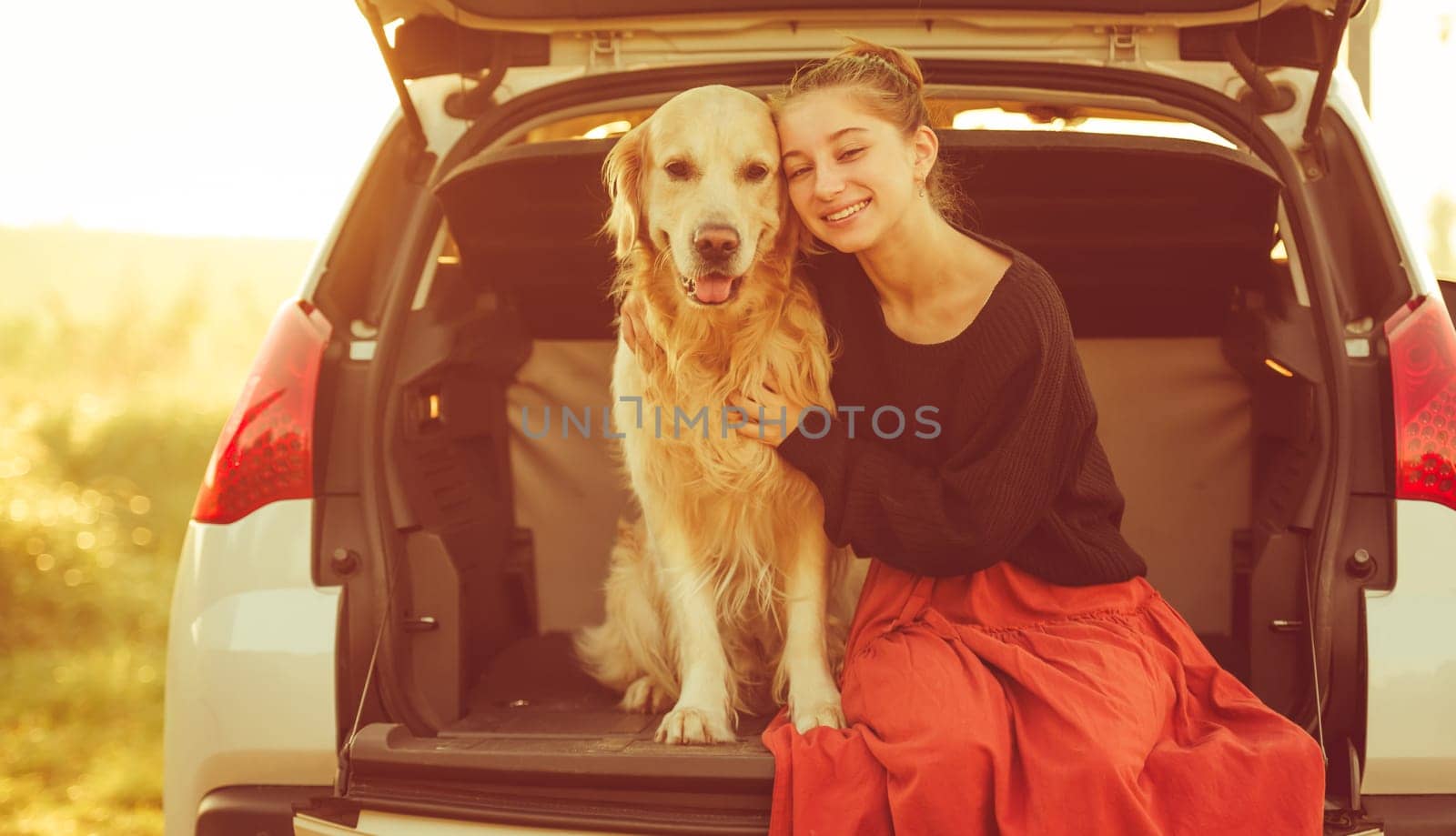 Beautiful girl with golden retriever dog sitting in car trunk and smiling. Pretty teenager hugging purebred pet doggy in vehicle at autumn nature