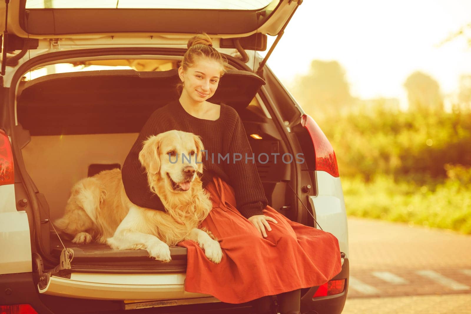 Beautiful girl with golden retriever dog sitting in car trunk and smiling. Pretty teenager hugging purebred pet doggy in vehicle at autumn nature