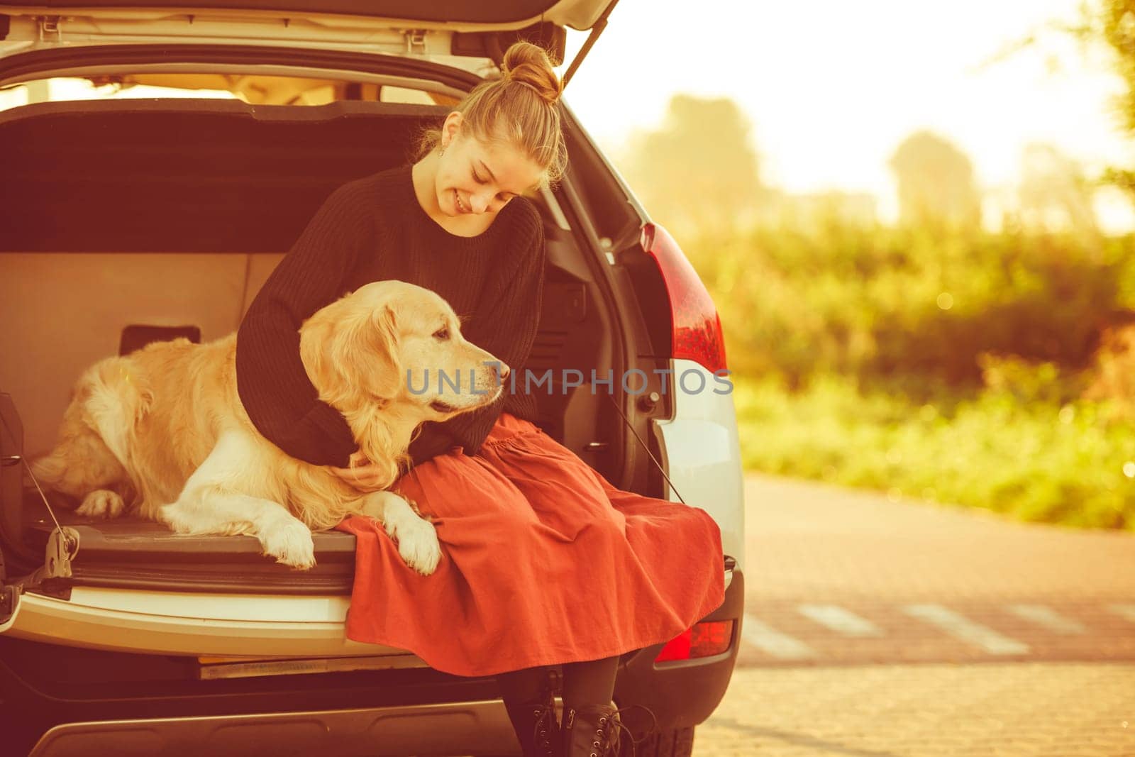 Beautiful girl with golden retriever dog sitting in car trunk and smiling. Pretty teenager hugging purebred pet doggy in vehicle at autumn nature