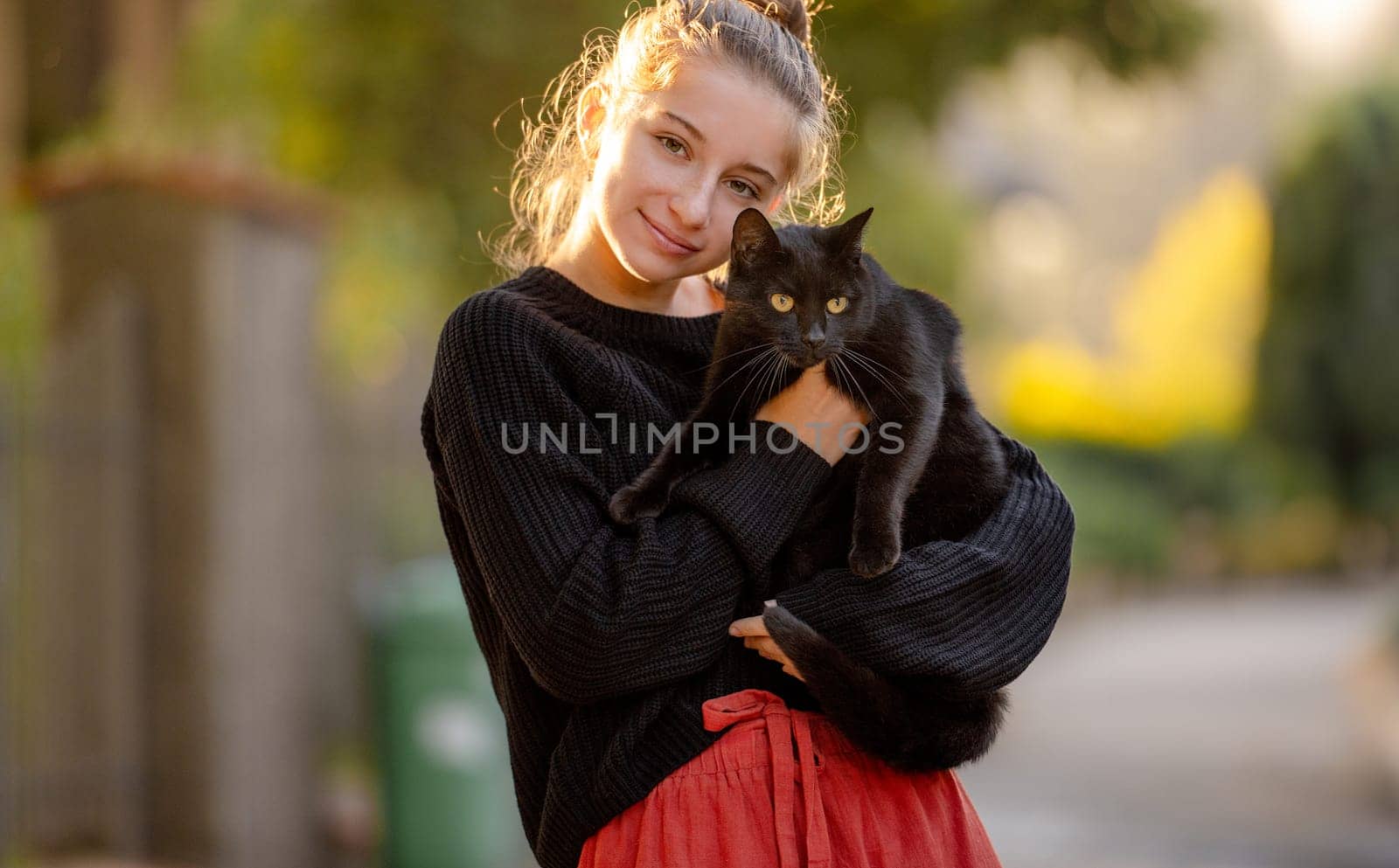Pretty girl hugging black cat outdoors at street with autumn light. Beautiful model teenager holding feline animal at park