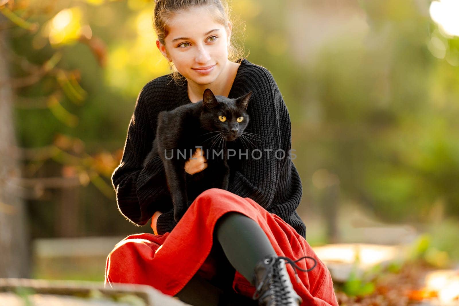 Pretty girl in red skirt hugging black cat outdoors at street with autumn leaves. Beautiful model teenager sitting with feline animal at park