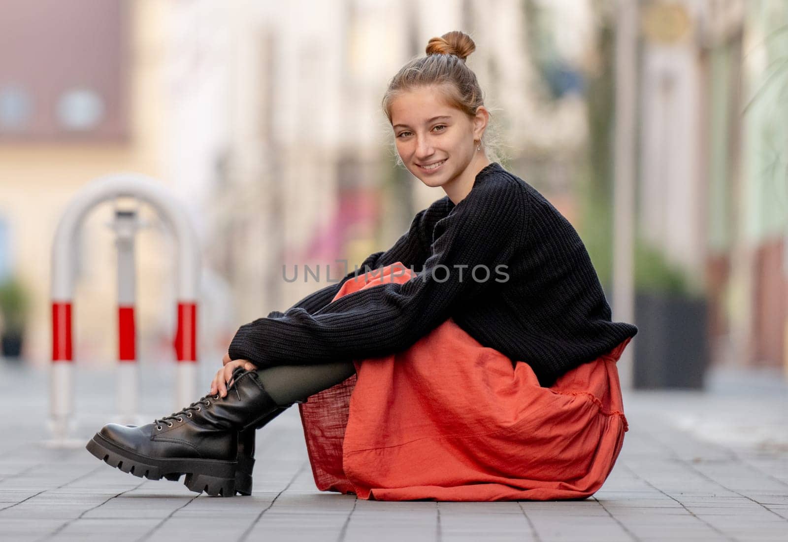 Beautiful teenager girl in red skirt sitting outdoors at street. Pretty teen model posing in trendy clothes