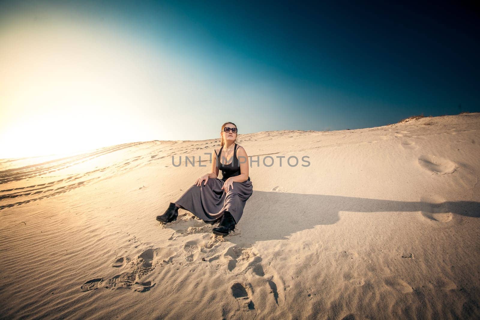 Beautiful woman in long skirt sitting on sand dune against blue sky and sun