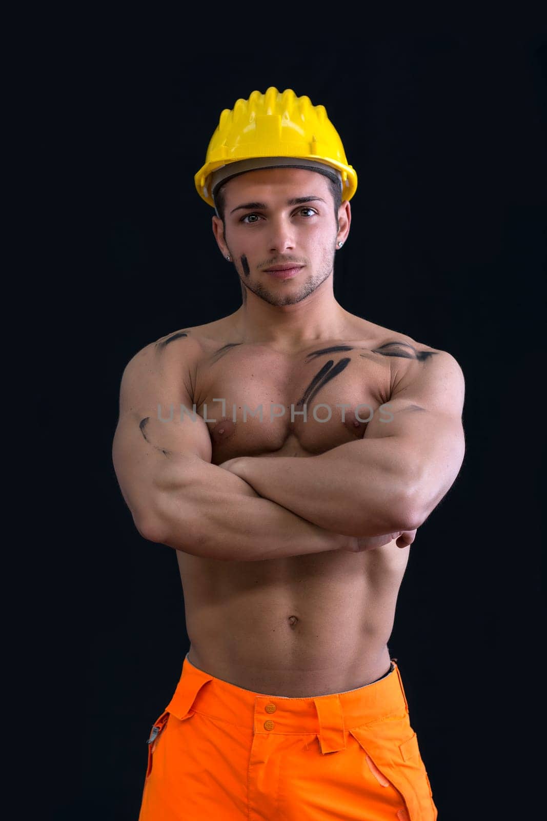 Muscular young construction worker shirtless wearing hardhat, arms crossed, isolated on black background