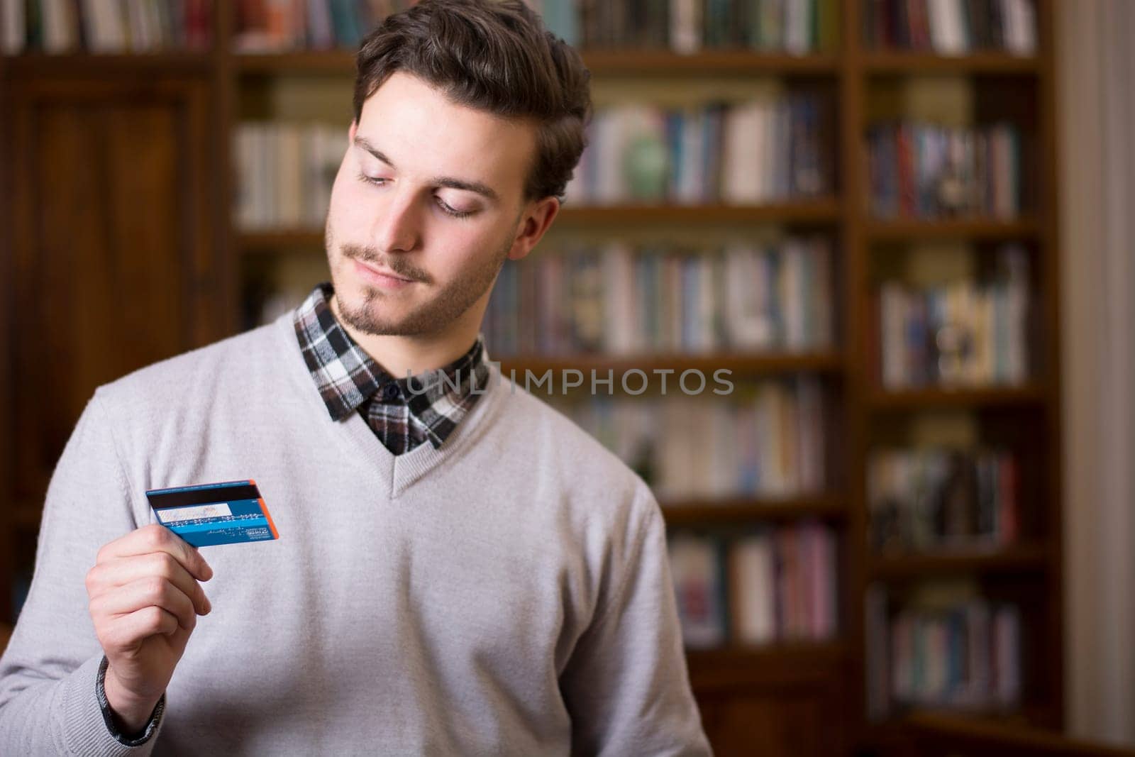 Attractive young man at home holding credit card, looking at it. Large copyspace