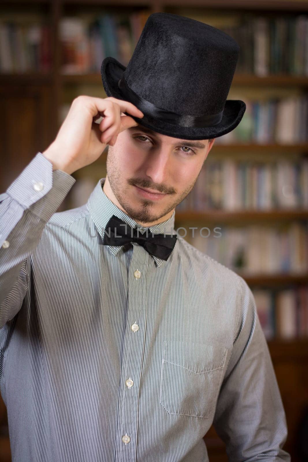 Attractive young man wearing top hat and bow tie, looking at camera. Indoors shot