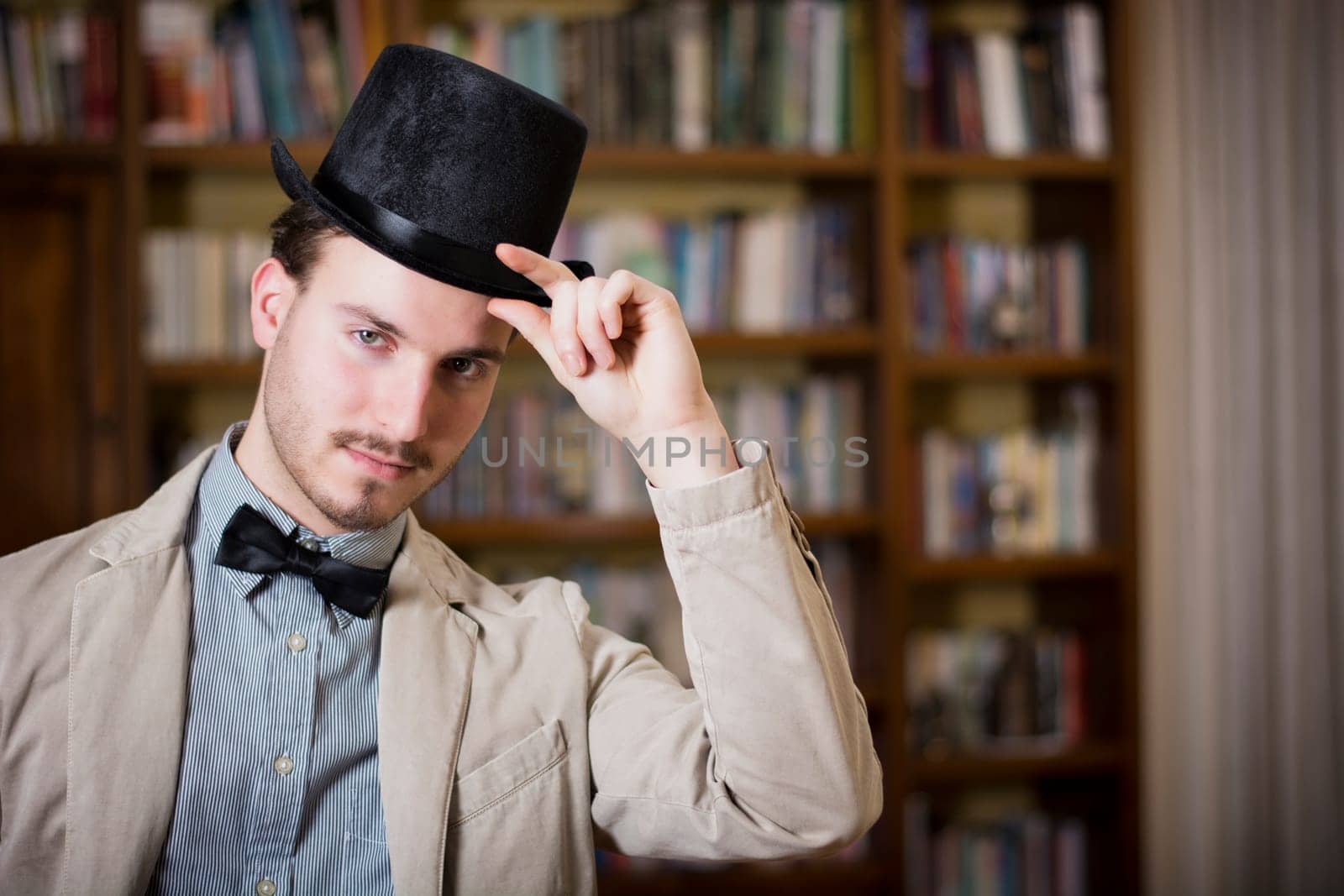 Attractive young man wearing top hat and bow tie, looking at camera. Indoors shot