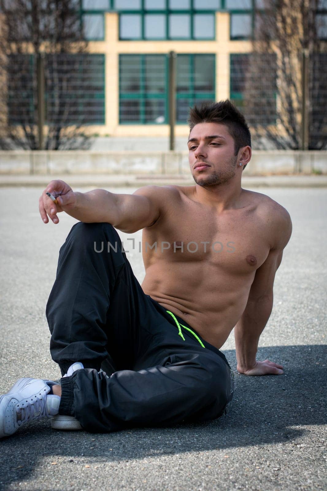 Shirtless muscular young man smoking cigarette outside, sitting on the ground